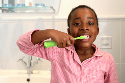 A child in a pink checkered pajama top brushes their teeth with a green electric toothbrush in a bathroom with white tiled walls and a sink.