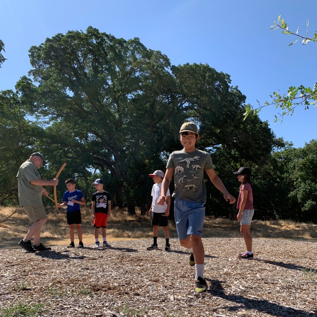 Five kids outside on a mulch path. Four of them are looking at their instructor, holding ancient hunting tools. One student is walking towards the camera, smiling.