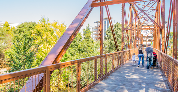 View from a rust-colored metal pedestrian bridge surrounded by green trees. Several people, including an adult pushing a stroller and two children, walk along the wooden pathway. The bridge’s beams form geometric patterns, and modern buildings are visible through the trees in the background under a clear sky.