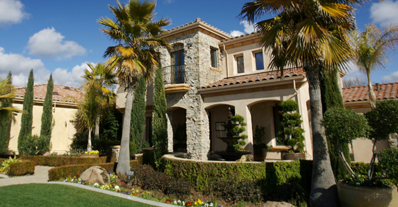 A two‑story house with a stone and stucco exterior, surrounded by tall palm trees, trimmed shrubs, and landscaped flower beds. The home features a small balcony, arched entryway, and a manicured lawn under a bright sky with scattered clouds.