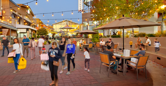 A lively outdoor shopping and dining area at dusk, with warm string lights hanging overhead. People stroll along a brick walkway while others sit at patio tables under umbrellas, eating and chatting. Shops and restaurants line both sides,