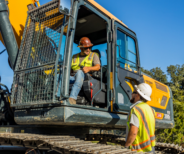 Construction workers in high-visibility vests and hard hats riding and standing beside a yellow excavator with a metal bucket on a sunny worksite.