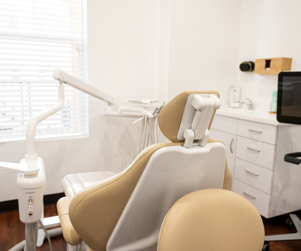 Dental chair with beige and white upholstery and an overhead light arm, set in a bright treatment room with cabinets and a computer monitor on the right.
