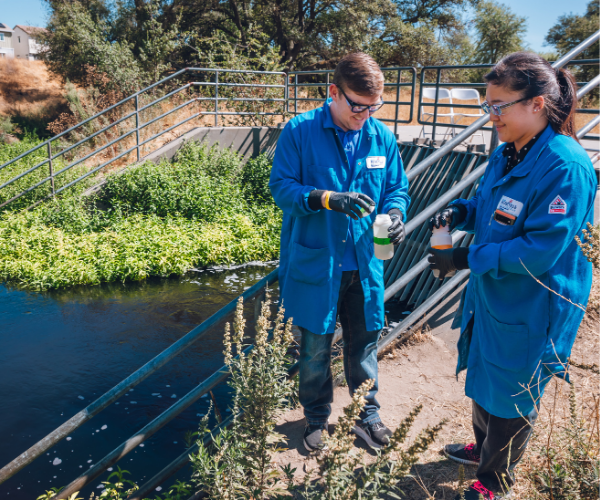 Two people in blue uniforms and gloves stand on a riverbank near metal stairs, inspecting and handling small plants by the water.