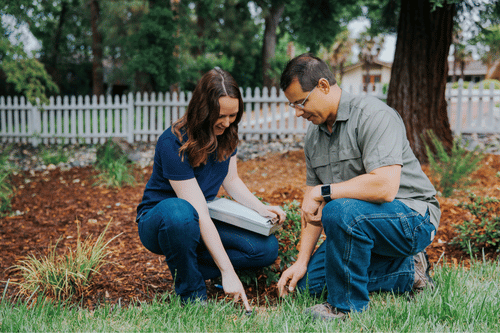 Two people kneeling in a yard, checking soil near plants and grass