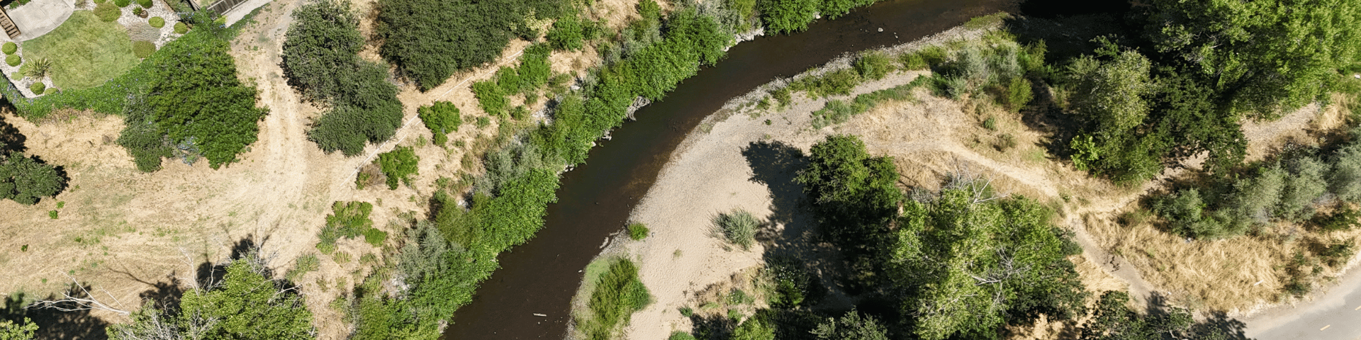 Aerial view of a winding creek flowing through a landscape with trees and houses on the left bank.