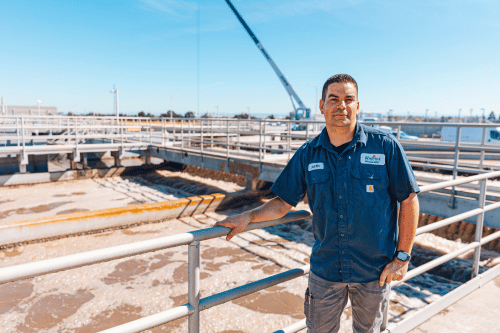 A man in a blue work shirt and gray pants stands on a walkway overlooking a large industrial water treatment facility with bubbling tanks and a crane in the background.