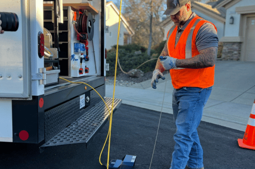 A utility worker in an orange vest uses a yellow cable to lower equipment into an open manhole. A service truck is parked nearby, and a traffic cone is visible on the right.