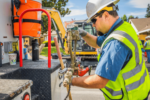 A worker in a hard hat and safety vest adjusts equipment on a utility truck, with a backhoe in the background.