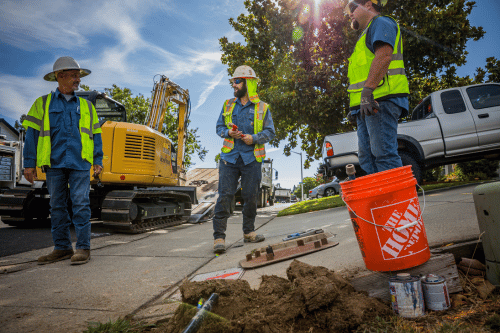Three construction workers in hard hats and safety vests stand around a construction site with an excavator and a truck in the background.