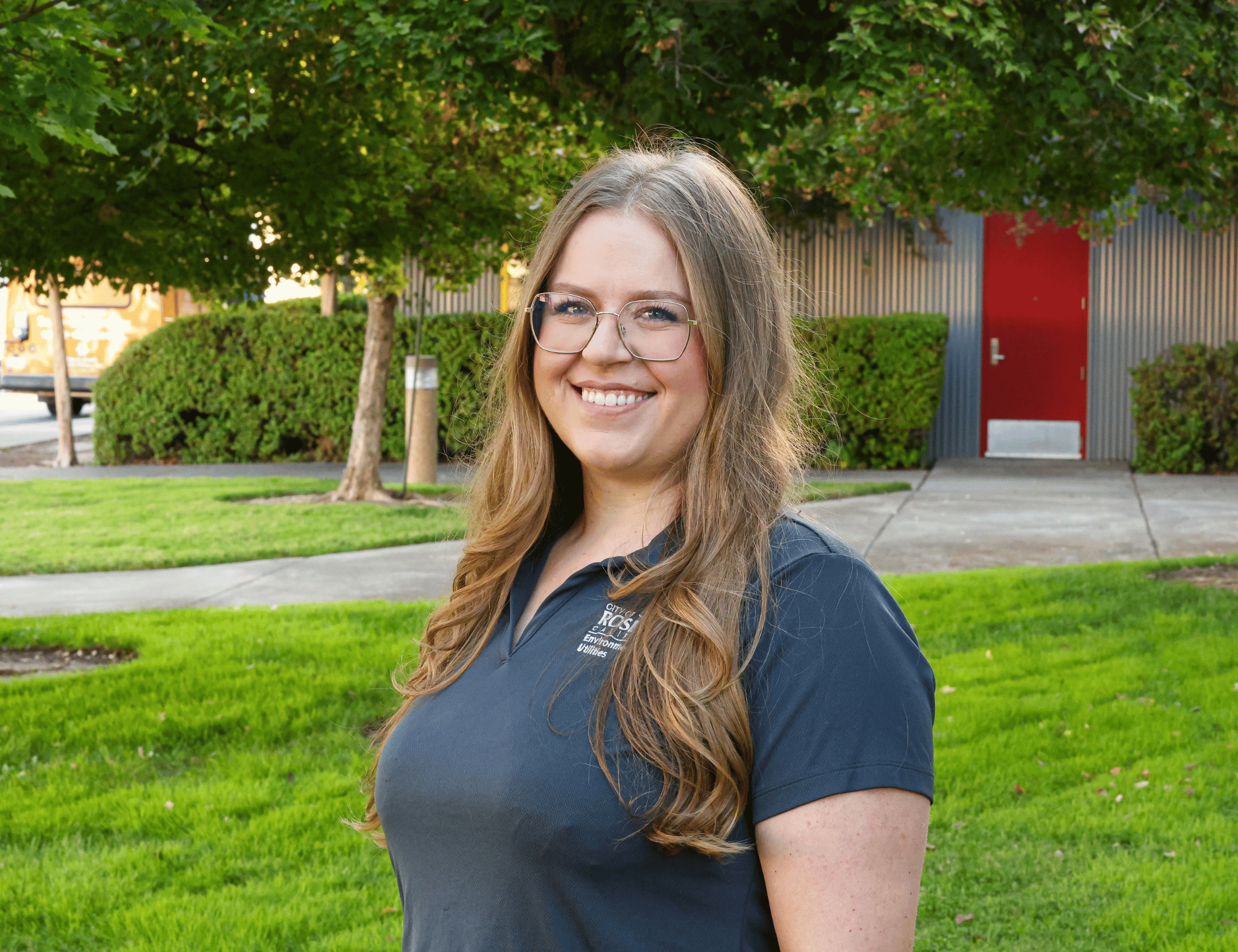 Alexa - Smiling person with long wavy hair and glasses standing on a green lawn in front of a building with red doors and trees