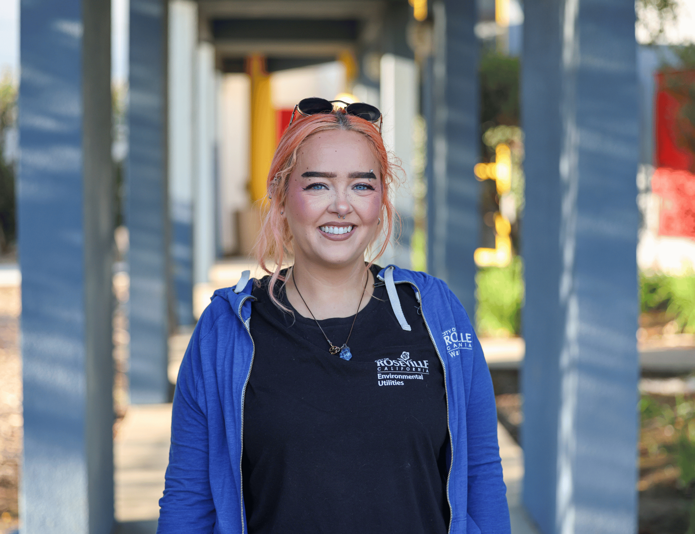 Brenna - Smiling person with reddish hair pulled back and sunglasses on their head, wearing a blue hoodie over a black T-shirt, standing on a sunny walkway between blue-painted columns