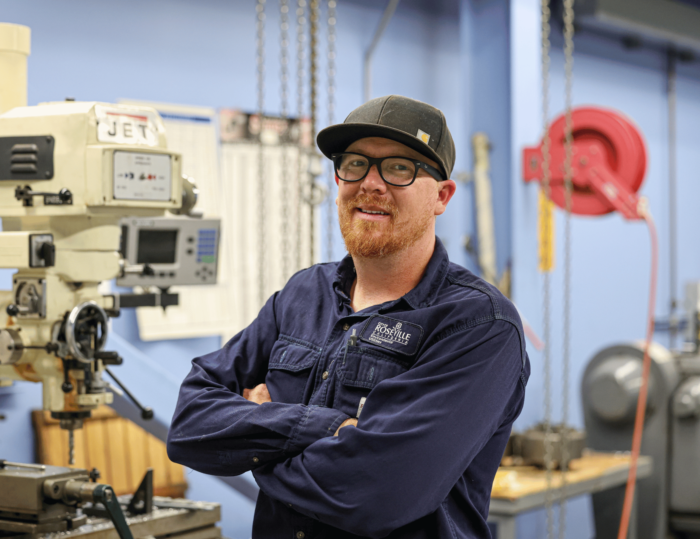 Zack - Worker in a dark jacket and cap stands with arms crossed in an industrial workshop, with a large machine, control panel, and hanging cables in the background