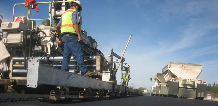 A construction worker stands on the rear of a large machine as it moves along a roadway.