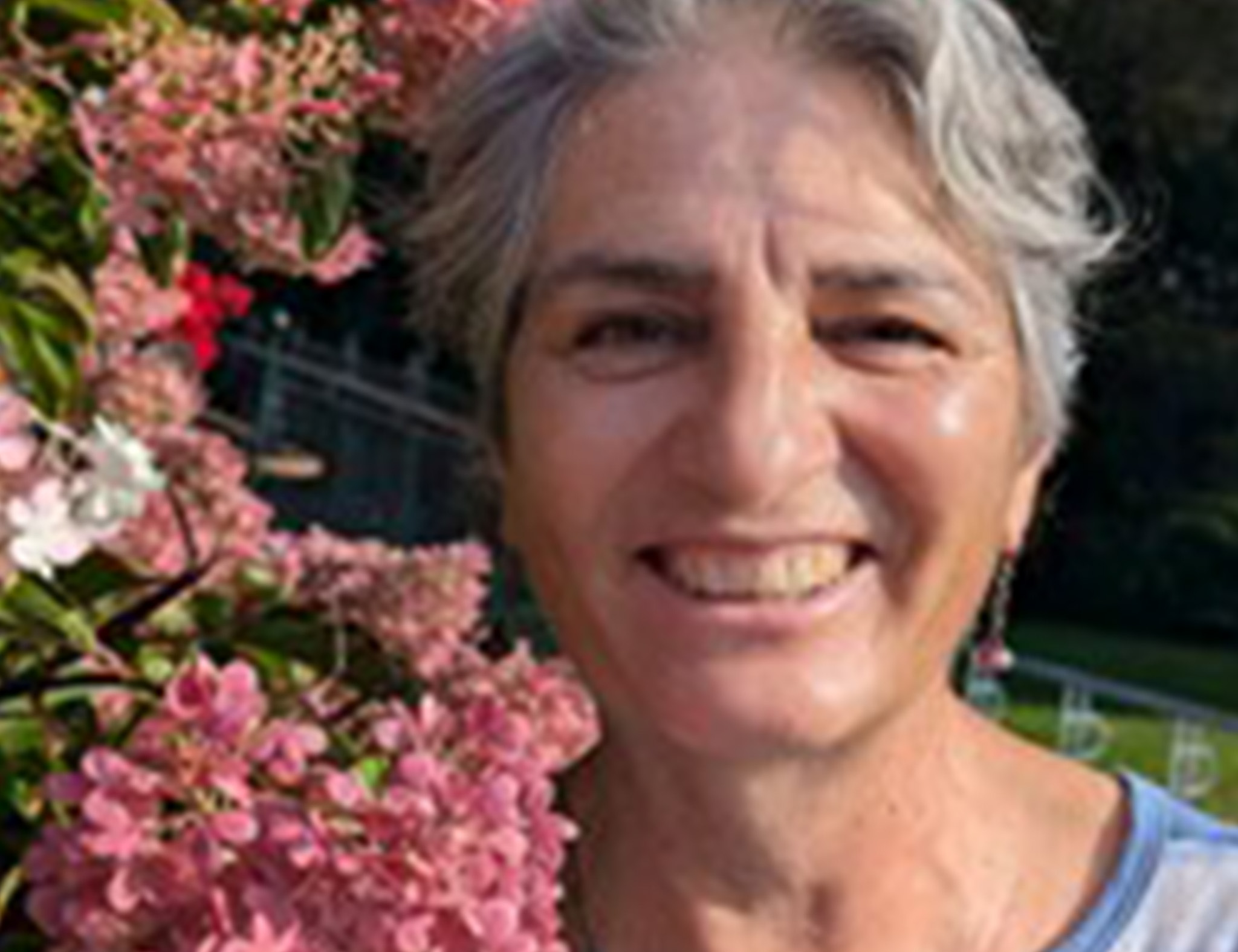 Close of older woman smiling as she stands next to pink flowers