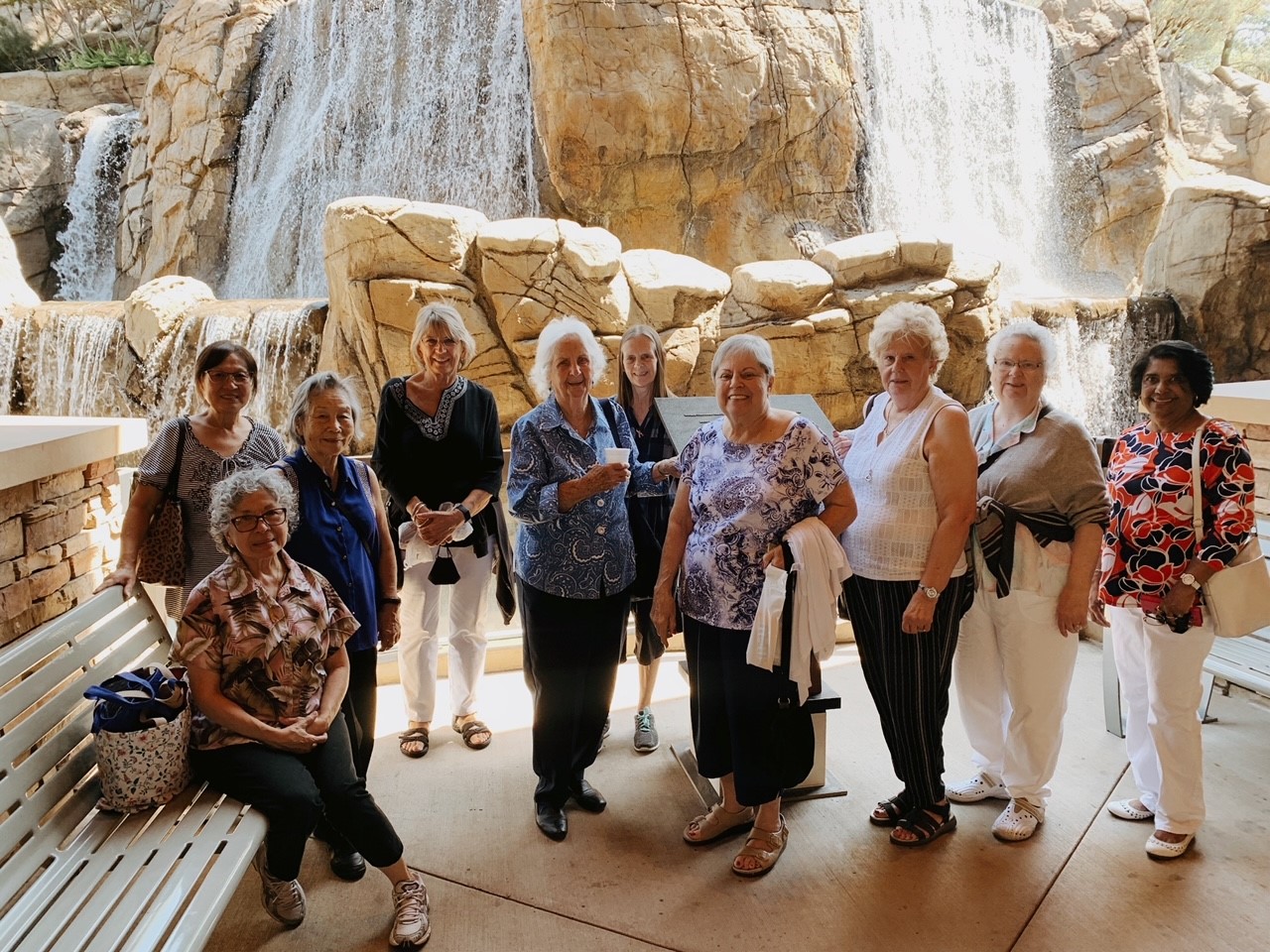 A group of 10 older adult women smiling in front of a rock and waterfall formation